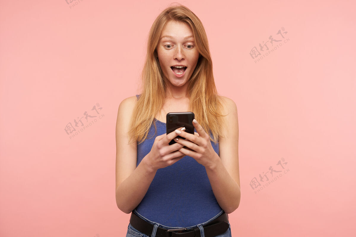 粉色年轻漂亮的长发红发女子穿着蓝色衬衫 一边兴奋地看着智能手机屏幕上的新闻 一边读着意料之外的新闻 隔离在粉色背景下电话提高衬衫