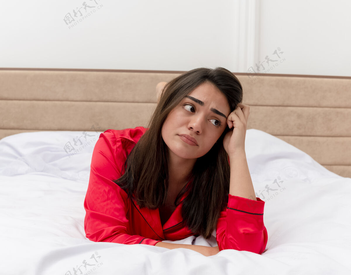 表情穿着红色睡衣的年轻美女躺在床上 脸上带着悲伤的表情 在家里的灯光背景下悲伤睡衣美丽