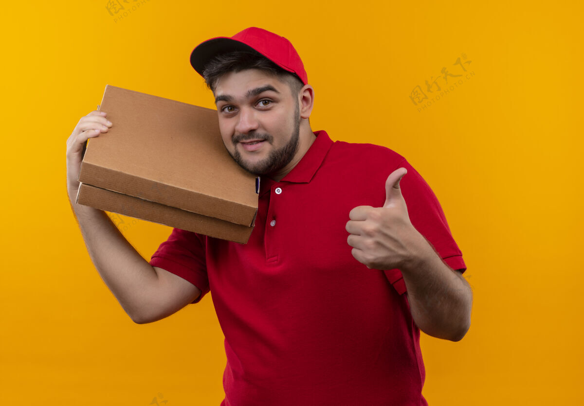 微笑年轻的送货员身穿红色制服 戴着帽子 手里拿着一叠披萨盒 微笑着竖起大拇指盒子拿着红色