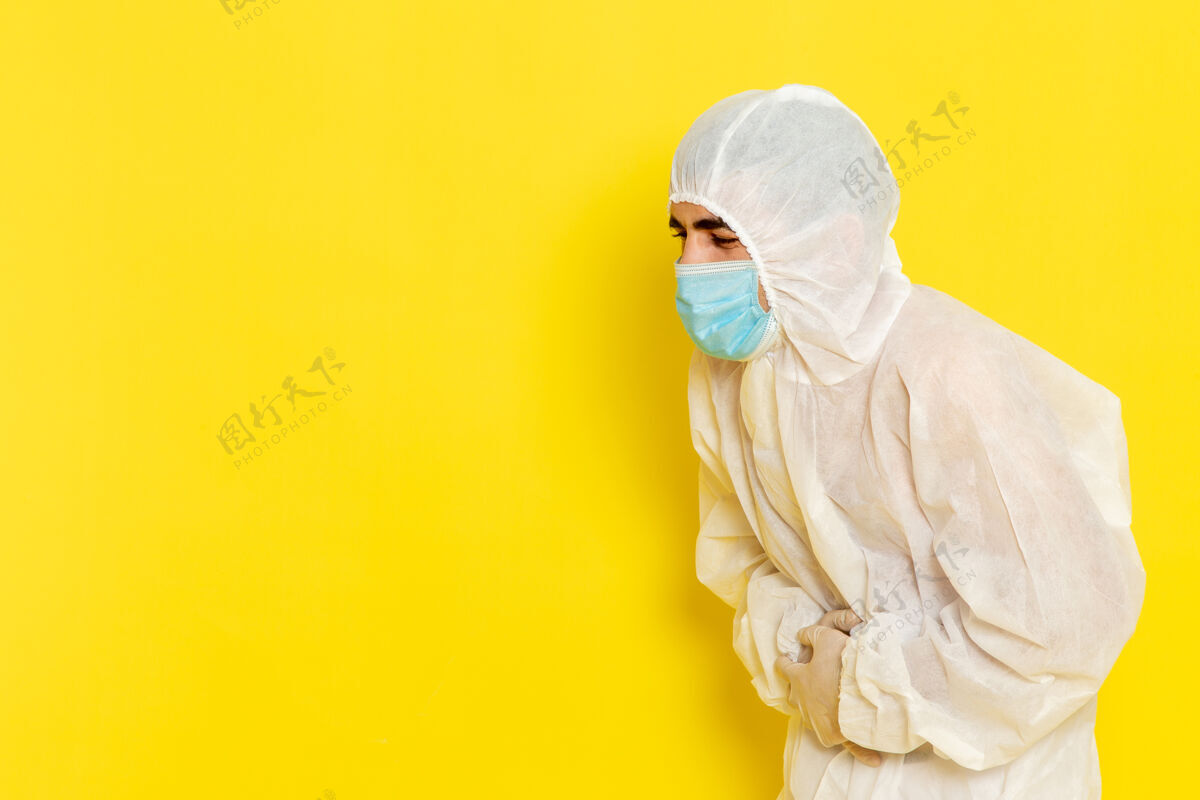 面具男性科学工作者的正面图 穿着特殊的白色防护服 戴着口罩 在浅黄色的办公桌上捂着肚子 科学工作者化学色危险照片光持有保护