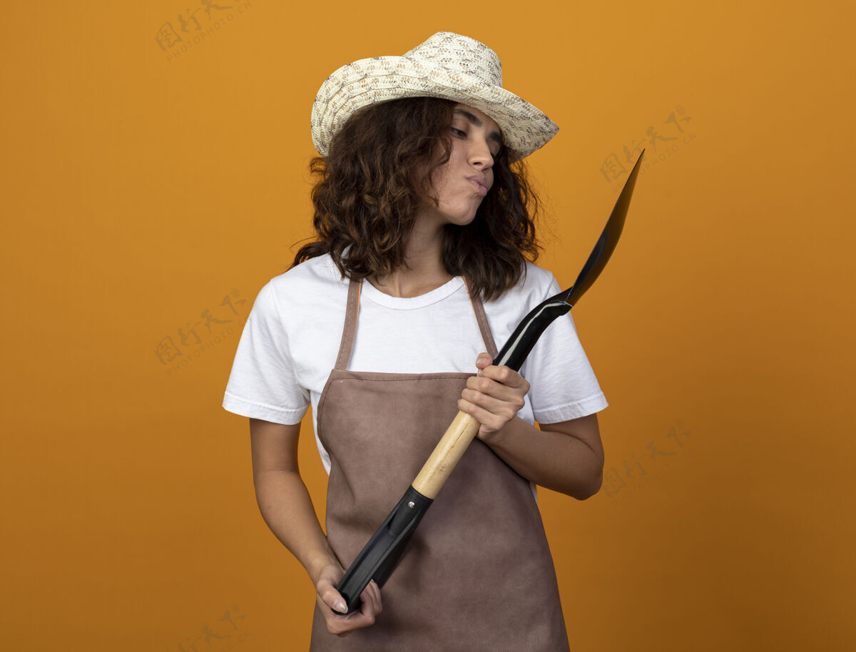 花园年轻的女园丁穿着制服 戴着园艺帽 拿着铲子 看着隔离在橙色墙上的铲子穿年轻黑桃