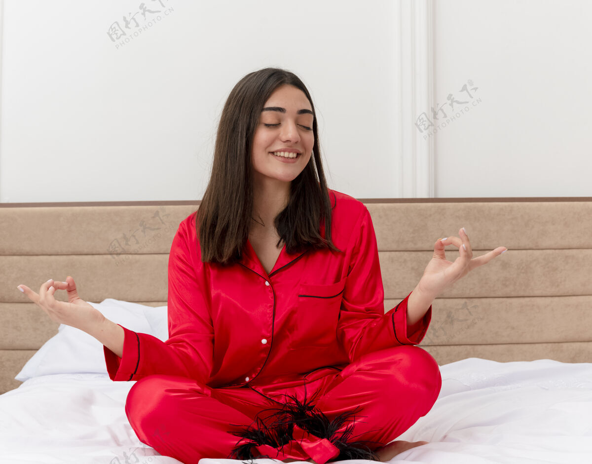 卧室穿着红色睡衣的年轻美女坐在床上 闭着眼睛放松 用手指在卧室内部的灯光背景下做冥想手势红色女人手势
