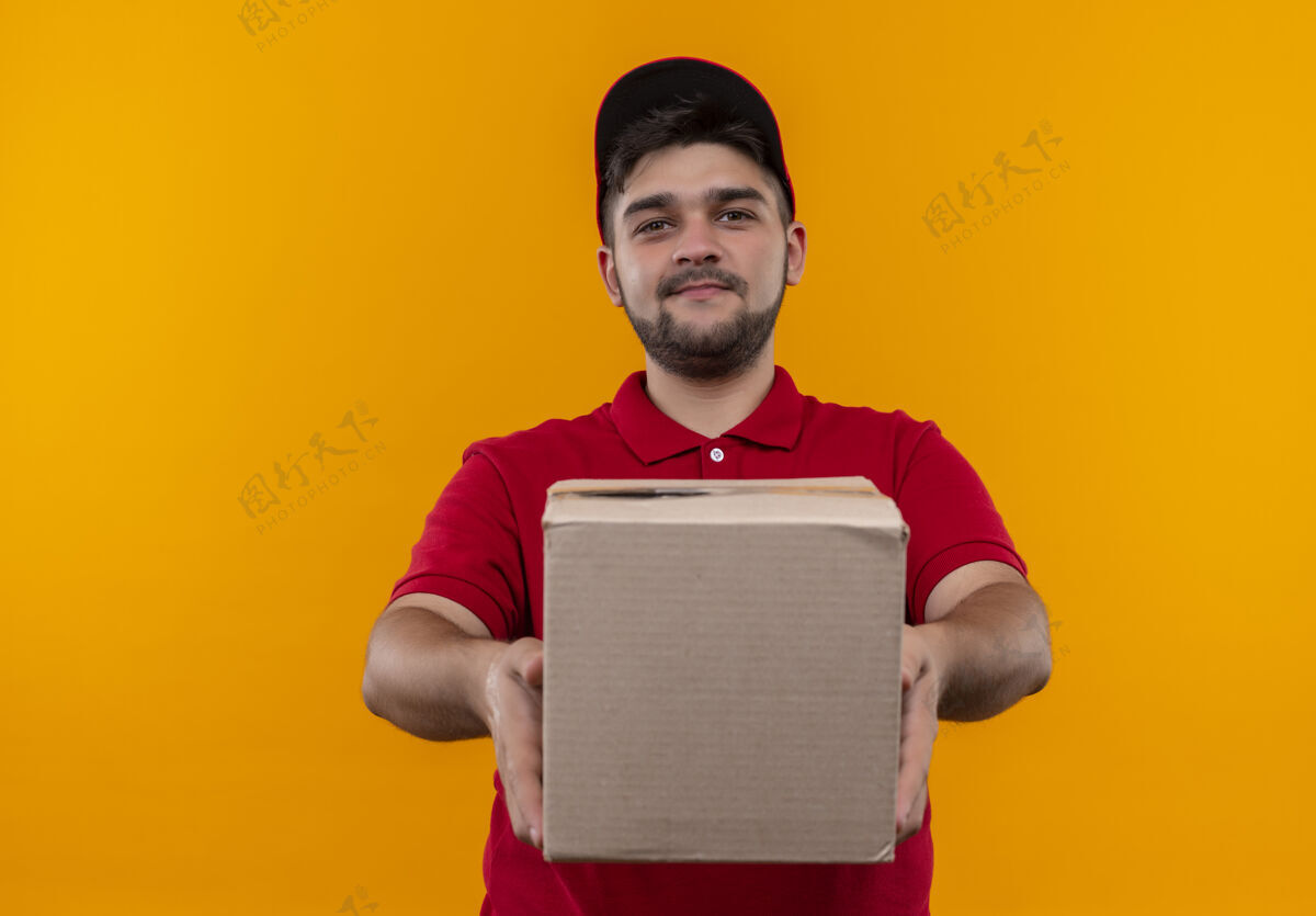 送货年轻的送货员穿着红色制服 戴着帽子 手里拿着盒子 微笑着自信拿着橙色包装