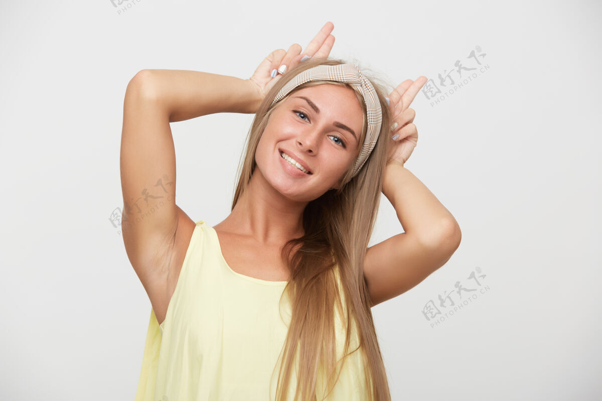 长迷人的蓝眼睛金发女性的肖像模仿耳朵举起双手 积极地看着相机 而在黄色衬衫的白色背景上摆姿势白色积极摆姿势