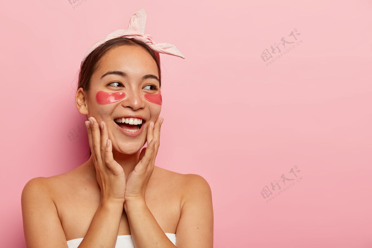 过程快乐的亚洲年轻女子戴着眼罩 开怀大笑 轻轻地抚摸着脸 看向一边 在水疗沙龙做美容 戴着粉色的头带 站在室内用毛巾包着高兴旁边模特