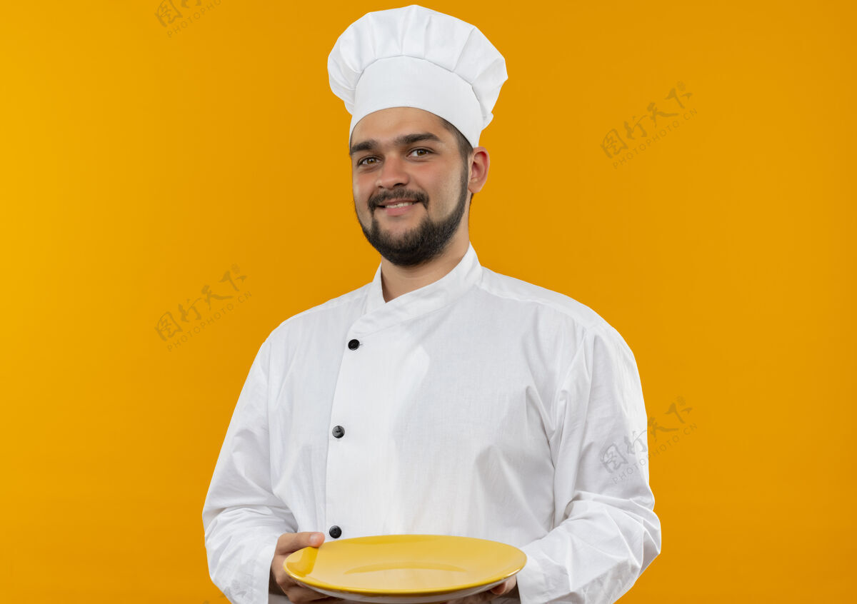 盘子微笑着的年轻男厨师身着厨师制服 手拿空盘子孤立在橙色的空间里年轻厨师拿着