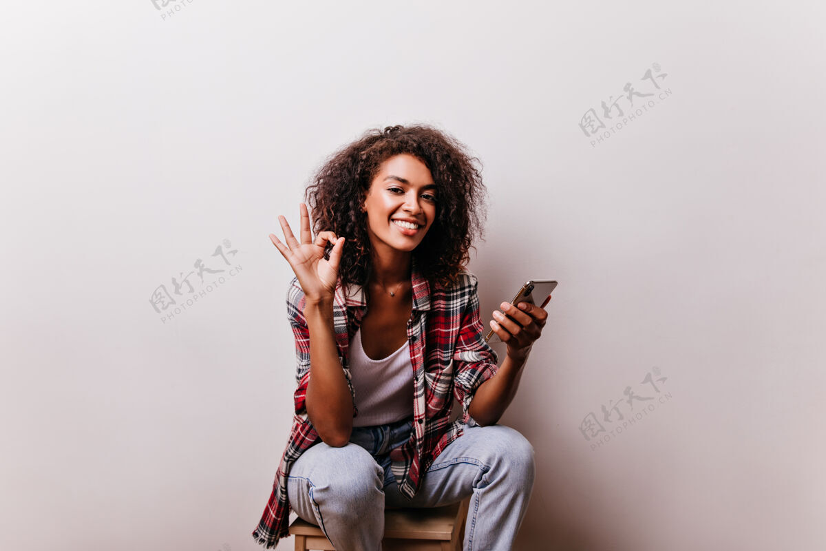 肖像迷人的非洲女孩摆出“好”的姿势卷曲的黑色女士拿着智能手机 带着浪漫的微笑年轻能源模型