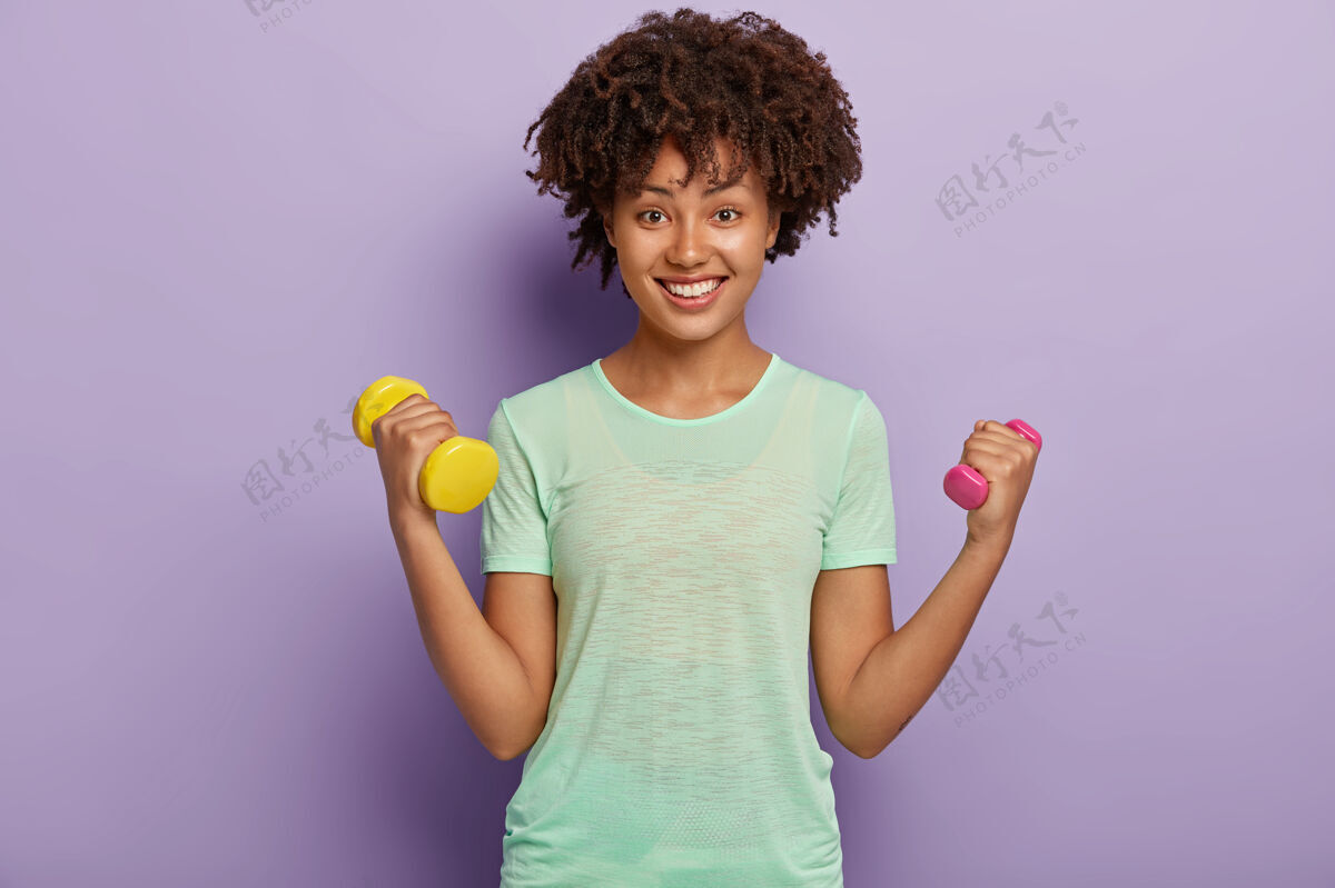 运动照片中可爱开朗坚强的女人举着哑铃双臂 训练肱二头肌 穿着休闲t恤 想要健康合身 露出愉快的牙齿微笑运动 女人的力量牙齿女性发型