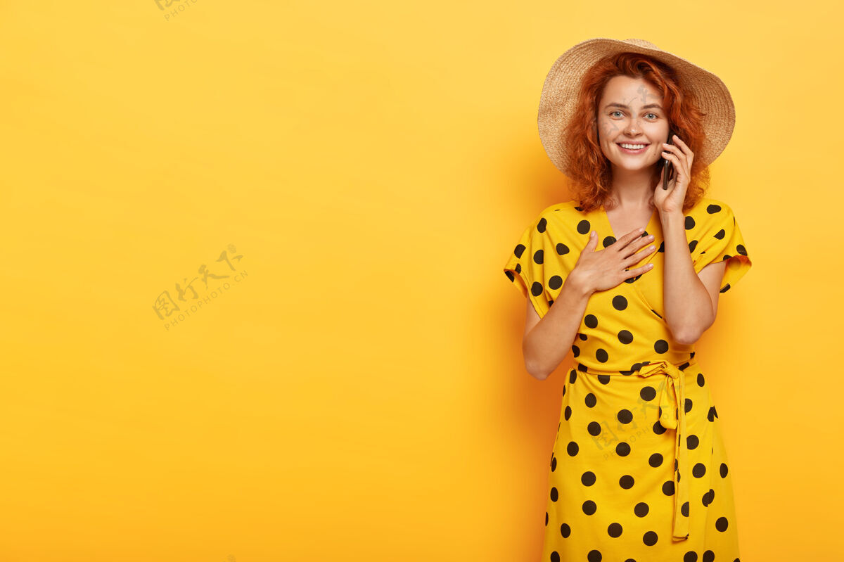 人容貌宜人 心满意足的年轻红发女子的画像 手掌放在胸前 通过智能手机听到令人心碎的故事 感觉印象深刻 穿着时尚的亮黄色夏装人模特电话