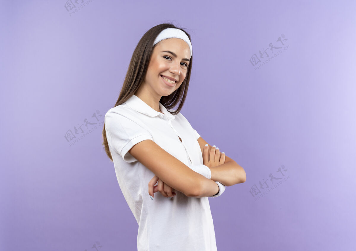 头带微笑的漂亮运动女孩戴着头带和腕带站在封闭的姿态孤立在紫色的空间站立佩戴微笑