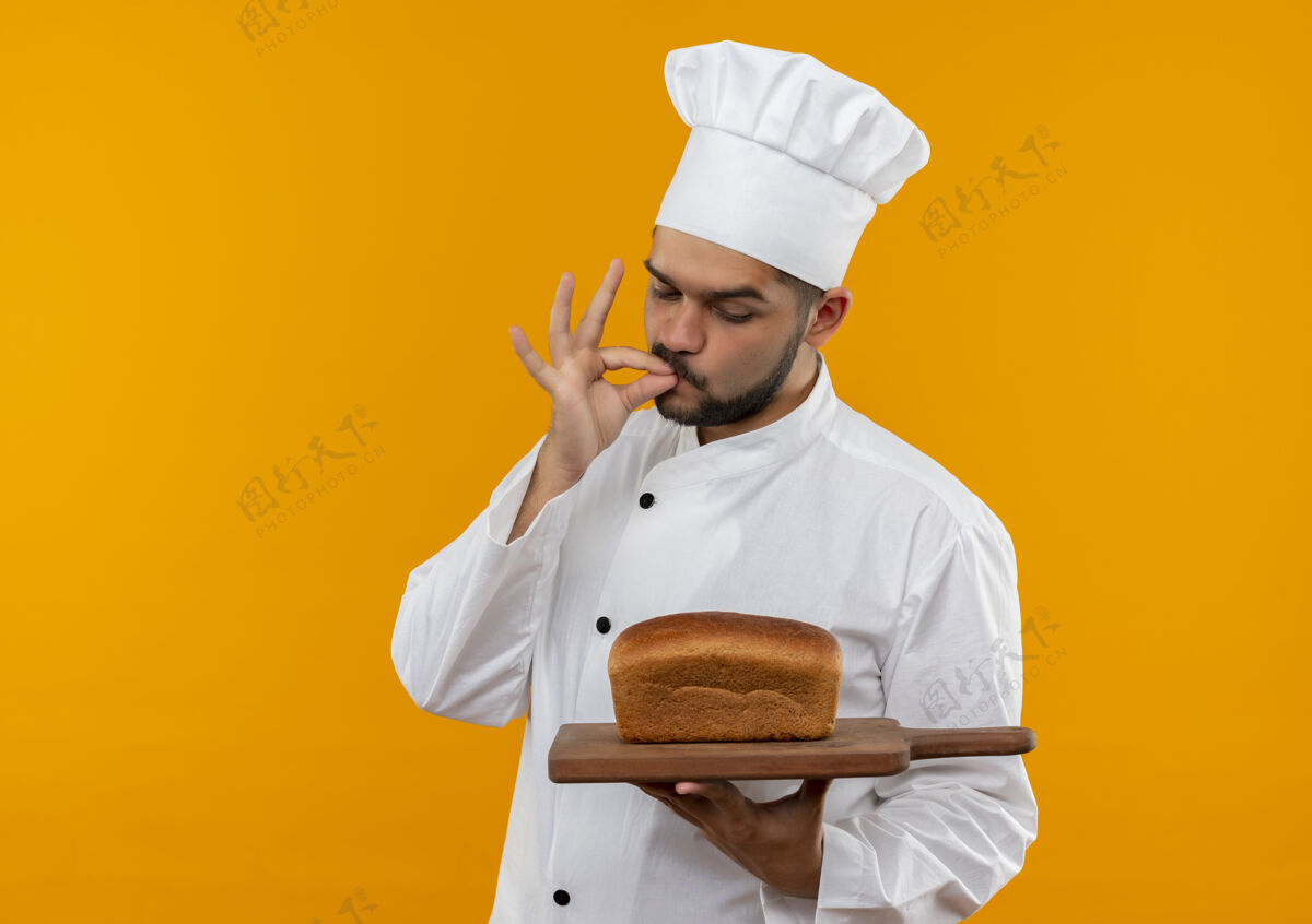 橘子身着厨师制服的年轻男厨师手拿着面包 看着砧板 做着美味的手势 孤立在橙色的空间里男性板拿着