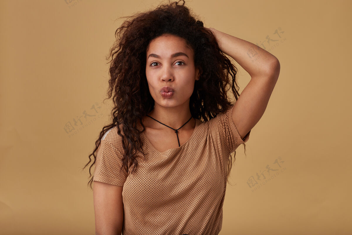 颈部积极向上的年轻可爱的棕色卷发女人 皮肤黝黑 噘着嘴唇 手放在脑后 孤立在米色上站立宽松休闲