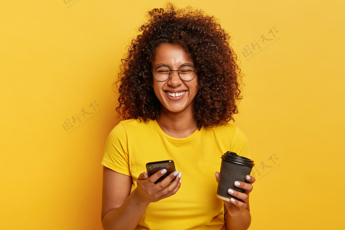 卷发过度活跃放松的黑发女性照片手持卡布奇诺外卖杯 在网上观看派对搞笑照片 使用现代电子设备 做视频会议 穿着随意信息眼镜美国
