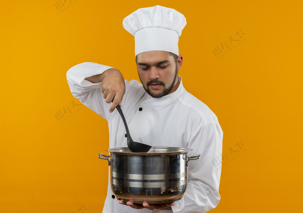 里面年轻的男厨师穿着厨师制服 拿着勺子 看着锅里面孤立的橙色空间看年轻厨师