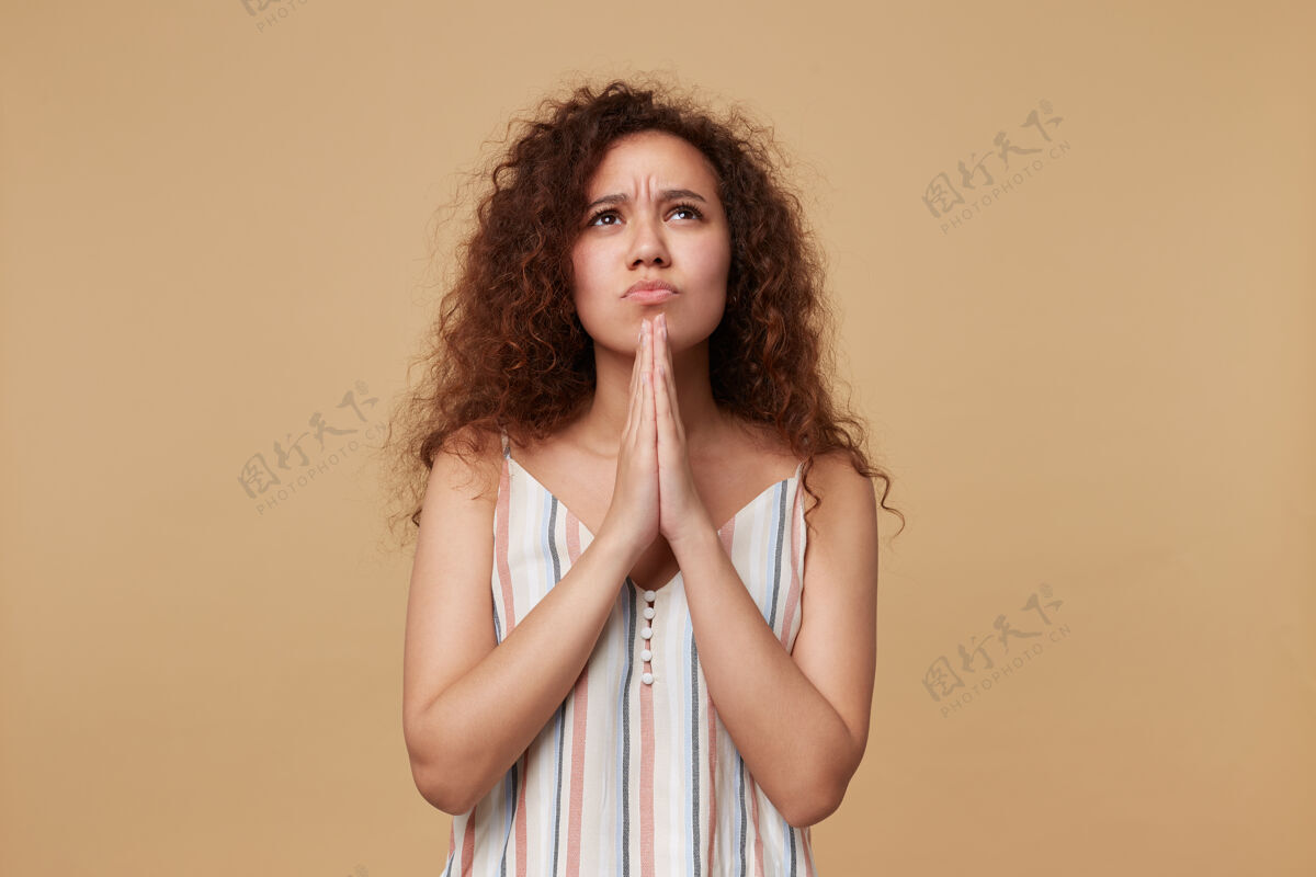 女士严肃的年轻卷发黑发女士 随意的发型 双手交叉祈祷 悲伤地向上看 孤立的米色女性摆姿势自然