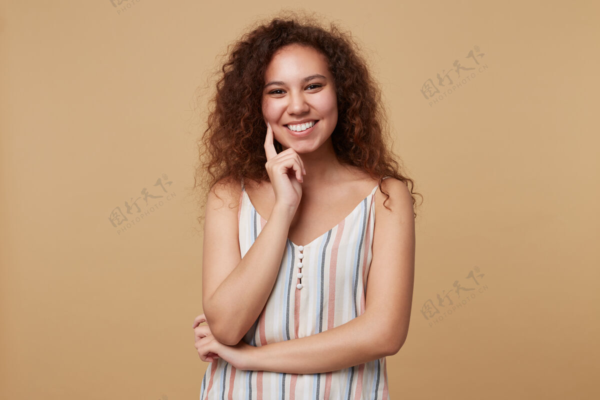 卷发年轻快乐的卷发黑发女士的横向肖像愉快地看着与广泛的微笑和触摸她的脸与举起的手 摆在米色长站着自然