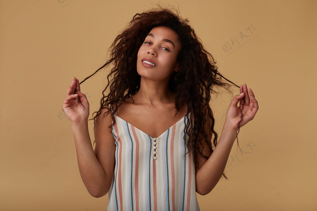 米色年轻可爱的正面长发卷曲的深色皮肤女人的画像拉着她的头发高兴的脸 摆在米色非洲女衬衫休闲