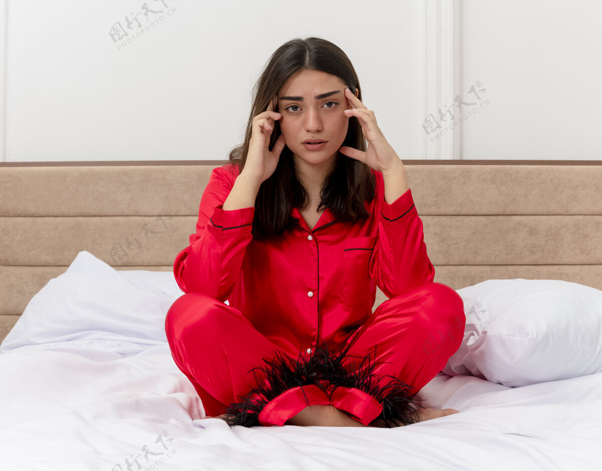 年轻穿着红色睡衣的年轻漂亮女人坐在床上 看着疲惫和无聊 在灯光背景下抚摸卧室内部的太阳穴太阳穴坐着无聊
