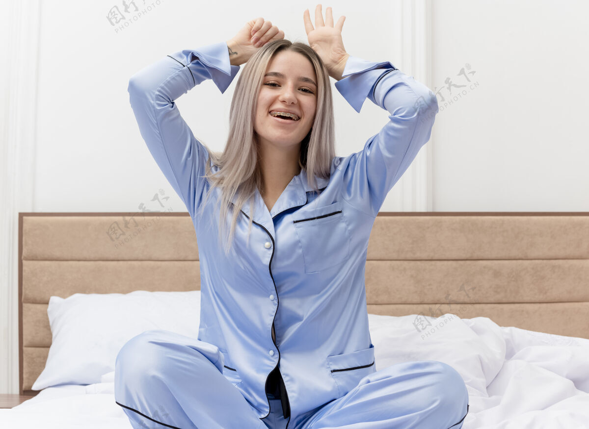 兔子穿着蓝色睡衣的有趣年轻漂亮女人坐在床上快乐欢快地模仿兔子耳朵在卧室内部浅色背景下微笑卧室坐着睡衣