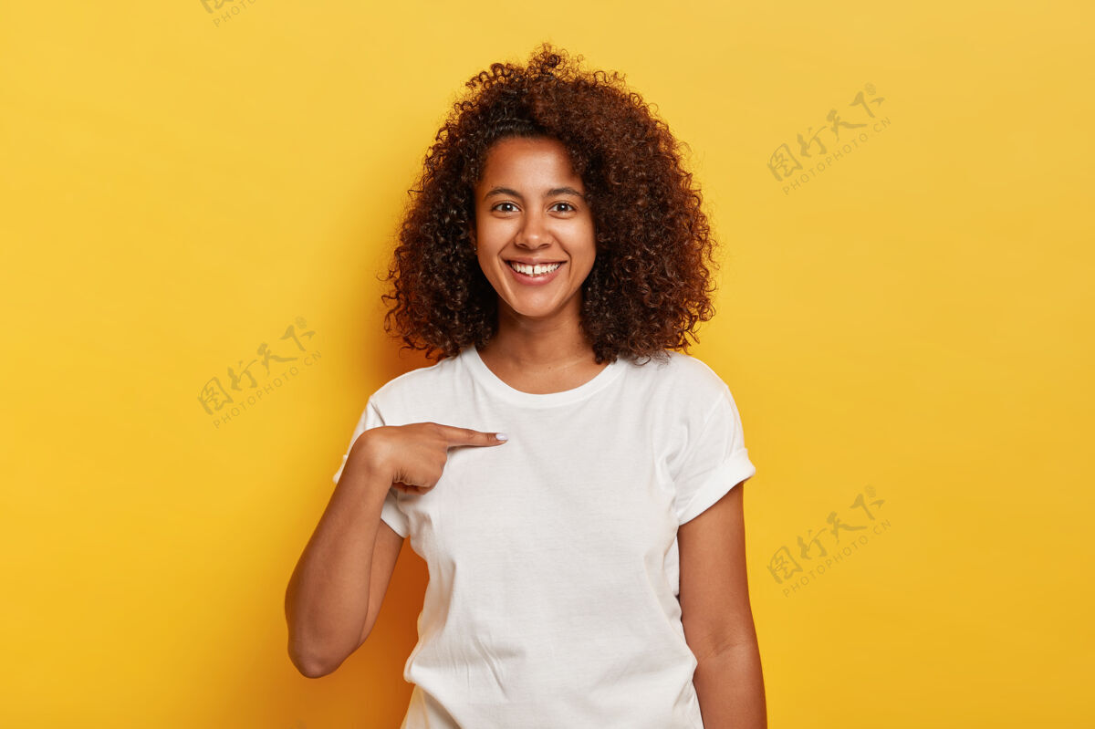 人笑容可掬的黑皮肤女孩指着自己 在白色t恤上展示了自己的模型空间 高兴地被选中 模特靠着黄色的墙壁无忧无虑的高兴的年轻非洲女人问我是谁乐观满意指向