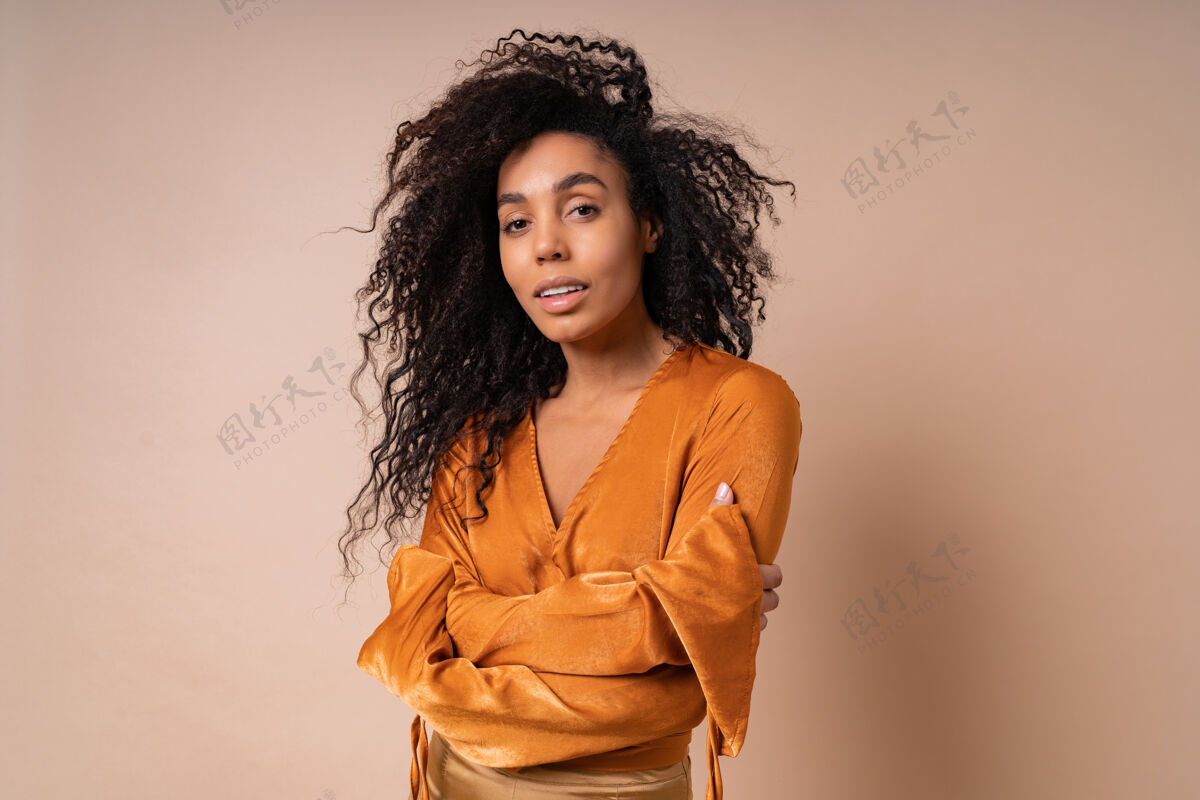 发型优雅的女士穿着橙色上衣 完美的皮肤 在米色的墙上摆姿势高跟鞋惊人的波浪形头发时尚非洲特写