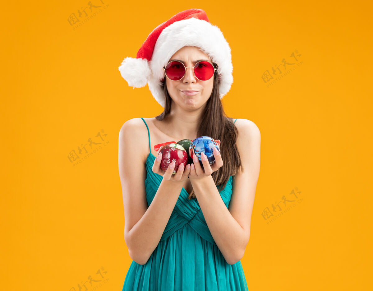 圣诞快乐戴着太阳眼镜 戴着圣诞帽 拿着玻璃球饰物的年轻白人女孩给人留下了深刻印象举行快乐装饰品