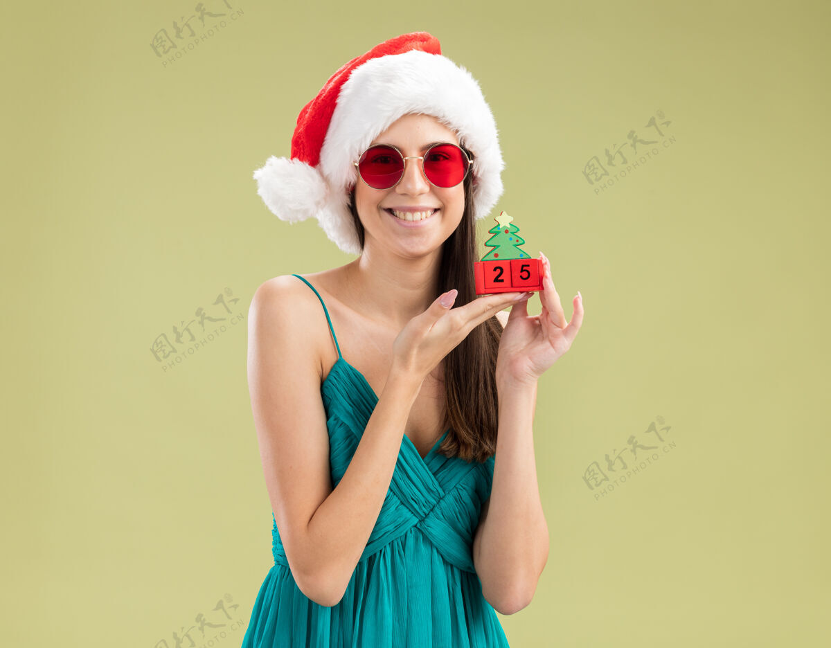 女孩戴着太阳眼镜 戴着圣诞帽 戴着圣诞树饰物的年轻白种人女孩高兴橄榄新