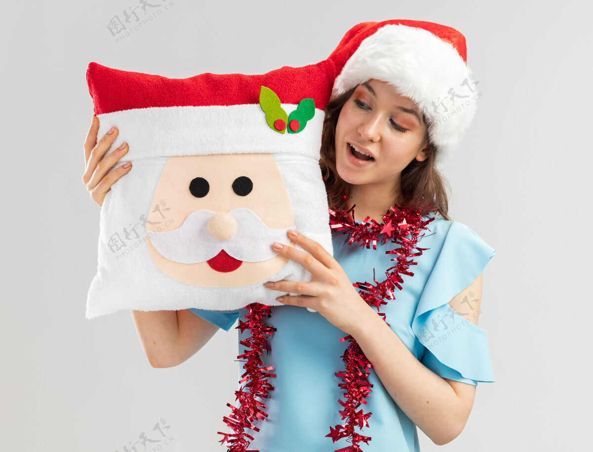 看一位身穿蓝色上衣 头戴圣诞帽 脖子上戴着金属丝的年轻女子 手拿圣诞枕头 面带微笑地看着它金属片新快乐