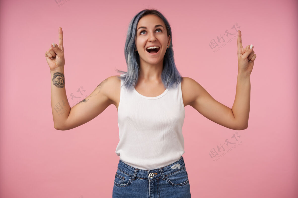 展示快乐的年轻纹身女子 短发竖起食指 站在粉红色的地面上 愉快地向上看发型站立短裤