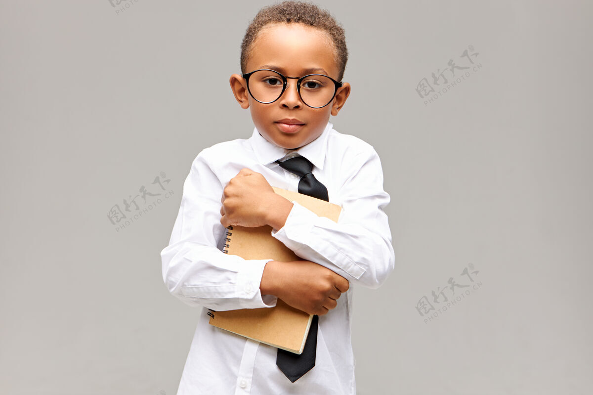 小一个英俊的深色皮肤 留着非洲短发的小学生的肖像 戴着眼镜 衬衫和领带 抱着抄写本 在新学校里感到害羞学习和知识领带抄写本非洲