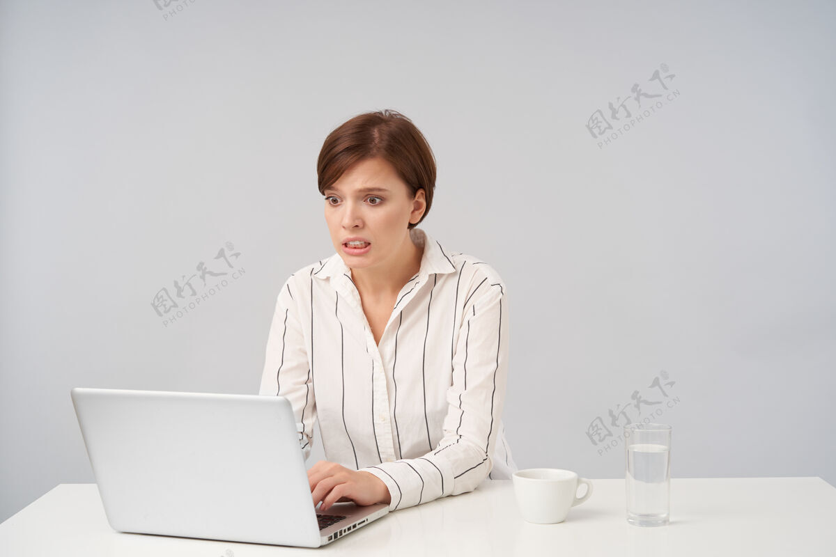 棕色年轻漂亮的棕色头发女性的室内肖像画 自然的妆容环绕着她的眼睛 露出牙齿 看着笔记本电脑的屏幕 困惑的表情 孤立在白色上尴尬困惑水