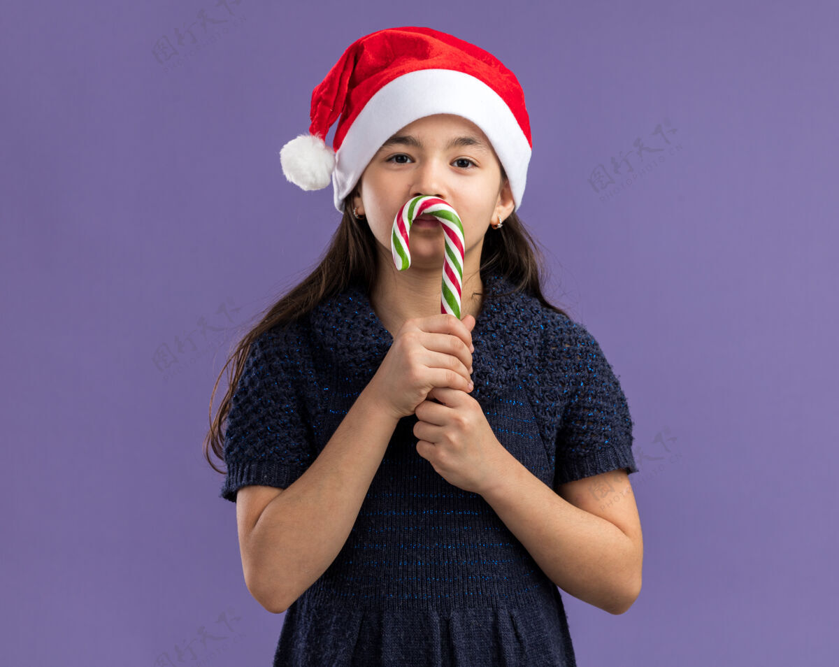 紫色小女孩穿着针织连衣裙 戴着圣诞帽 手里拿着糖果手杖 脸上洋溢着快乐和积极的笑容小站着圣诞