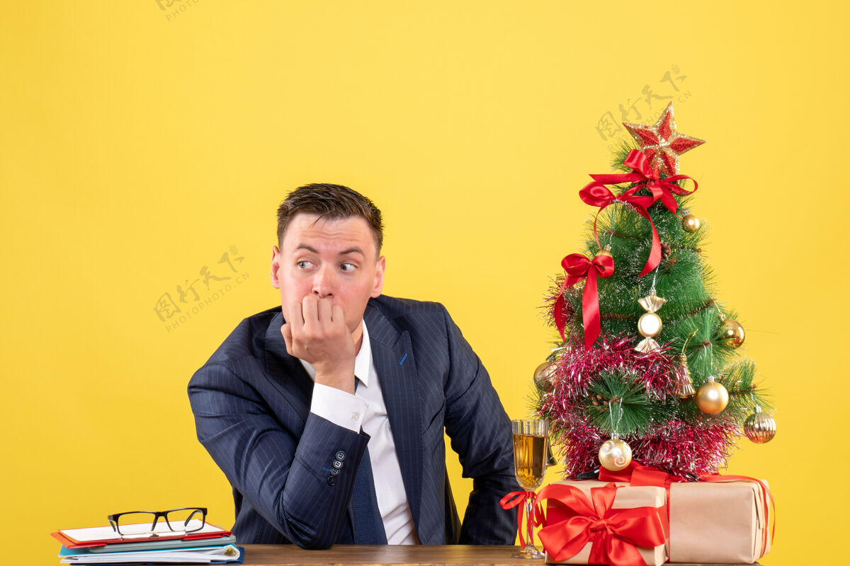 主任前视图：体贴的男人坐在圣诞树旁的桌子旁 黄色的礼物坐着树办公室