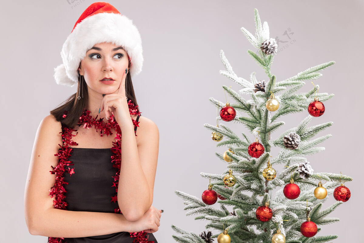 体贴体贴的年轻漂亮的白人女孩戴着圣诞帽 脖子上戴着金箔花环 站在装饰好的圣诞树旁 手放在下巴上 看着隔离在白色墙上的一面漂亮年轻圣诞节