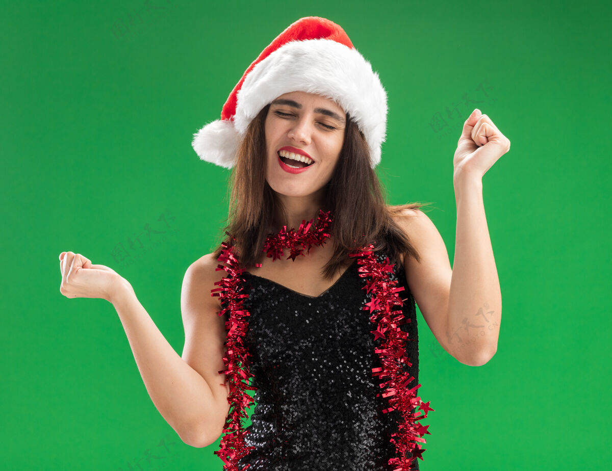关闭快乐与闭上眼睛年轻漂亮的女孩戴着圣诞帽 脖子上的花环显示是的姿态隔离在绿色的墙壁上脖子穿着帽子