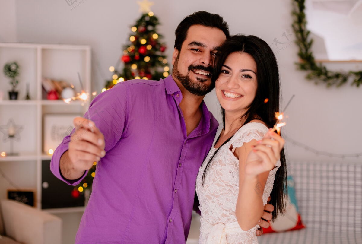 年轻年轻美丽的情侣 带着火花的男女 拥抱着幸福的爱情 在装饰好的房间里一起庆祝圣诞节 背景是圣诞树火花装饰一起
