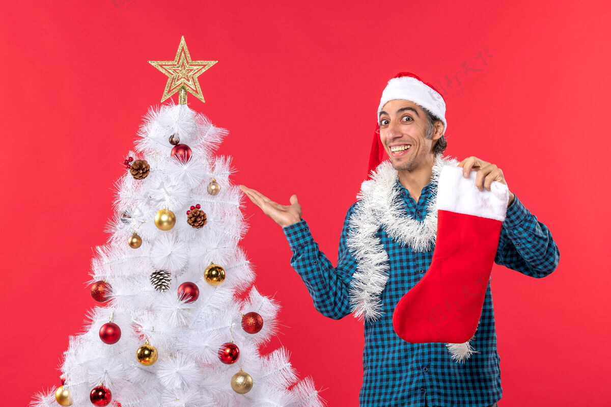 男人兴奋的年轻人戴着圣诞老人的帽子 穿着蓝色的衬衫 手里拿着圣诞袜子 指着红色的圣诞树圣诞老人年轻人帽子