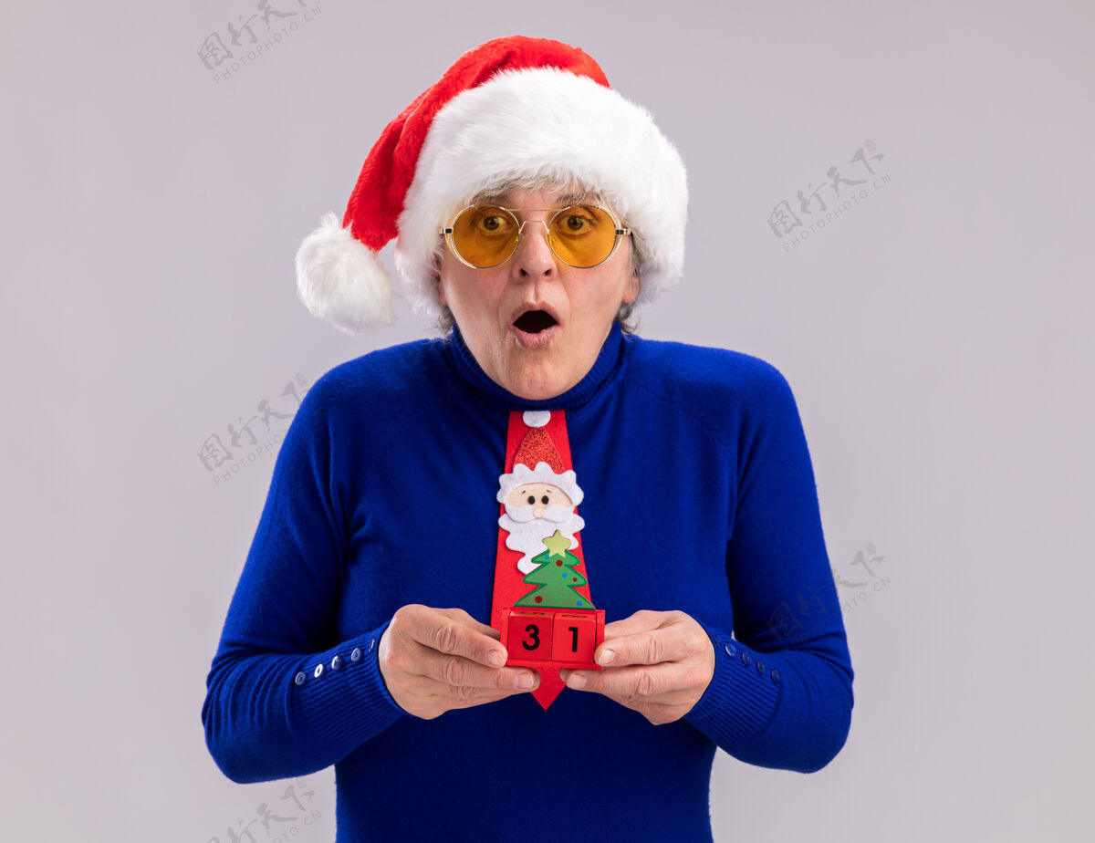 震惊戴着太阳眼镜 戴着圣诞帽 打着圣诞领带 手拿圣诞树饰物的老妇人惊呆了圣诞老人眼镜年