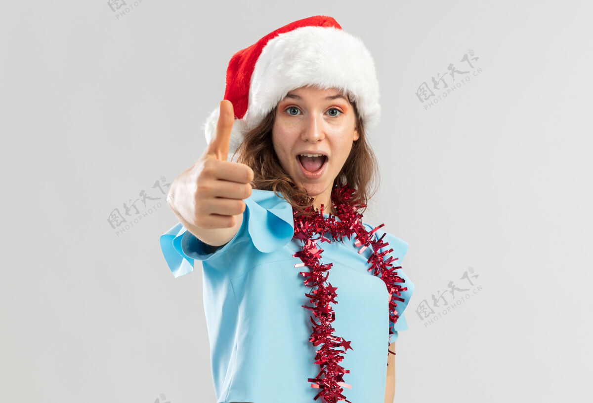 年轻一位身穿蓝色上衣 戴着圣诞老人帽 脖子上戴着金属箔的年轻女子 看上去既高兴又兴奋地竖起大拇指圣诞向上快乐