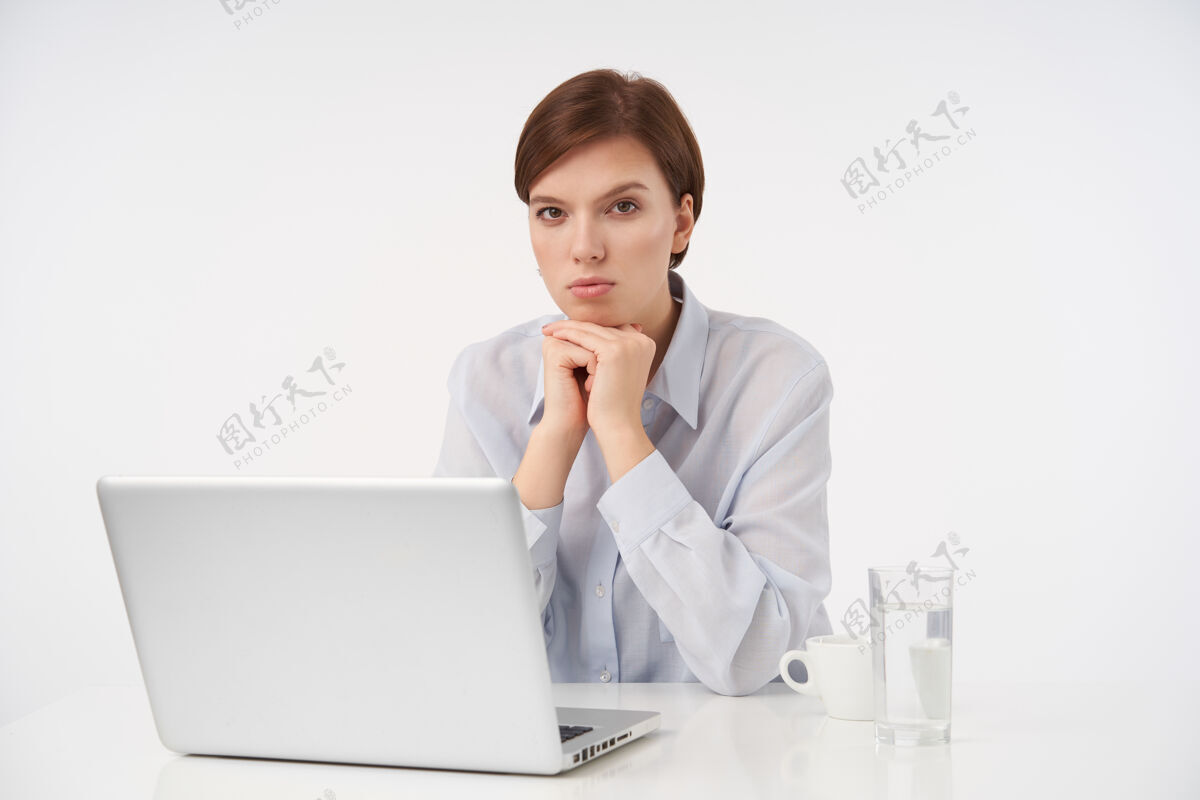 折叠严肃的年轻漂亮的棕色眼睛的黑发女性 短而时髦的发型 手放在下巴下面 表情严肃 摆出白色的姿势摆姿势笔记本电脑化妆品