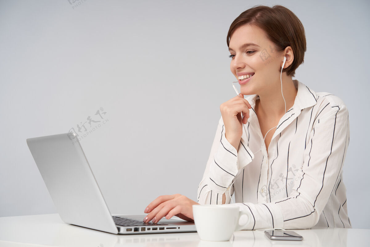 女士一个年轻的棕色头发的女人的肖像 她留着时髦的短发 手放在键盘上 坐在粉色的笔记本上 开心地微笑着 手举在下巴下面工作积极开朗