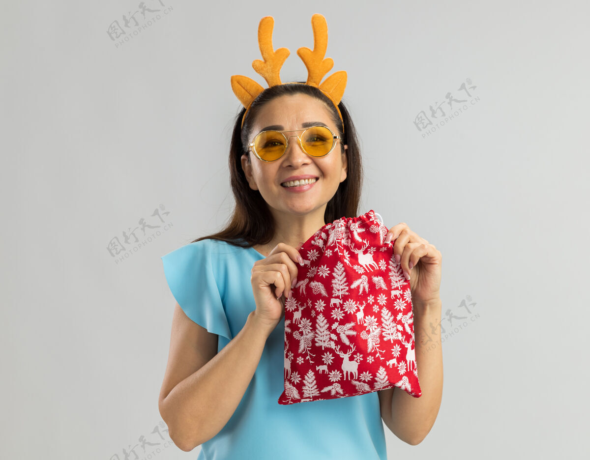 顶穿着蓝色上衣 戴着滑稽的鹿角边 戴着黄色眼镜 手里拿着圣诞礼物 脸上挂着大大的笑容的快乐年轻女子年蓝色圣诞快乐