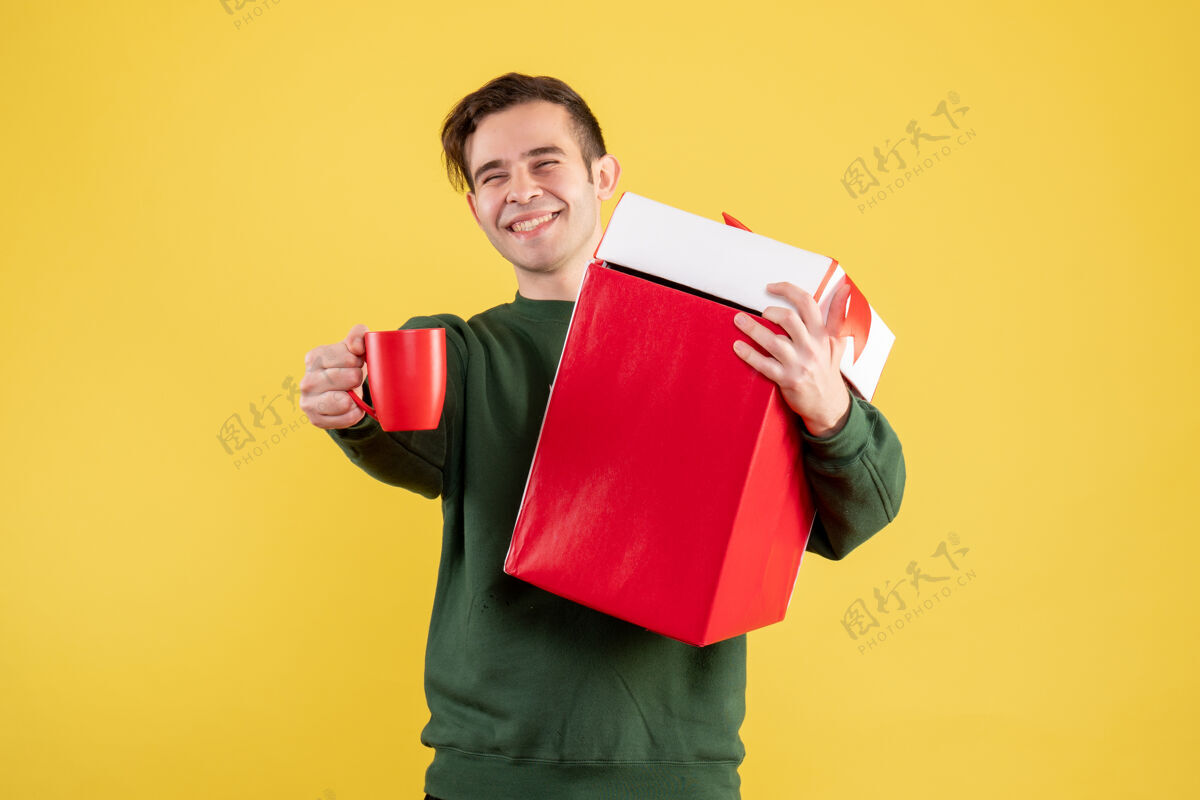大正面图：穿着绿色毛衣的年轻人手里拿着大礼物 红色的杯子站在黄色的椅子上人成人站着