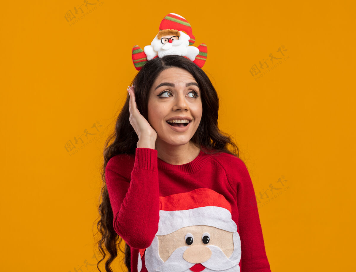 漂亮令人印象深刻的年轻漂亮女孩戴着圣诞老人的头带和毛衣看着一边保持手在头上橙色背景隔离圣诞老人印象深刻圣诞老人