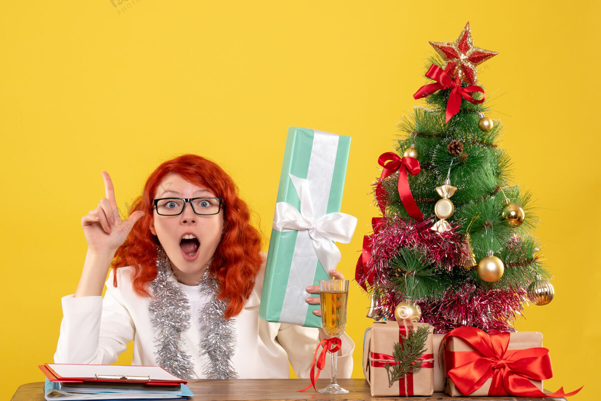 插花前视图：女医生坐在黄色背景上 手里拿着圣诞礼物和圣诞树花礼物前面