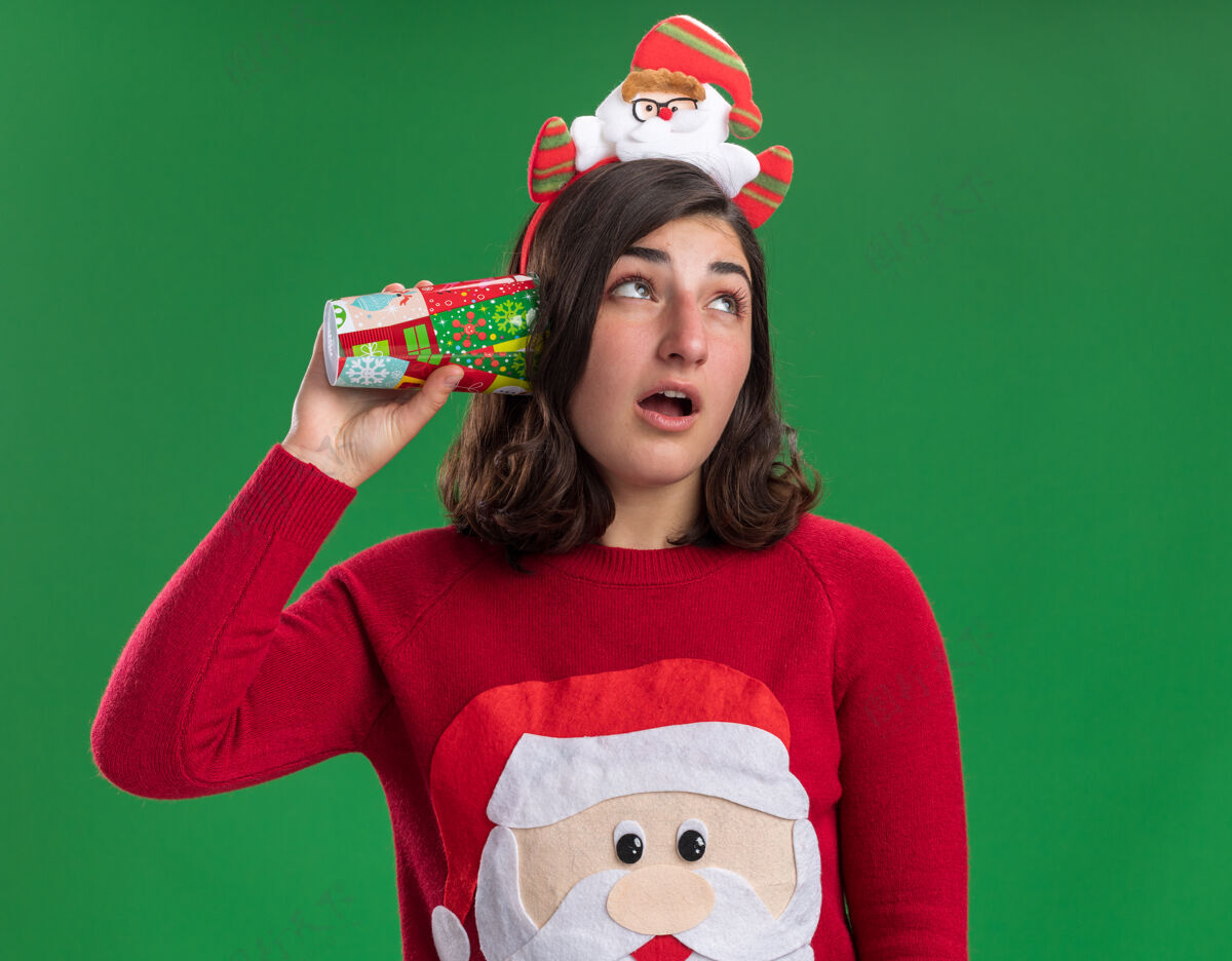 年轻人穿着圣诞毛衣 戴着圣诞帽的小女孩把五颜六色的纸杯捂在耳朵上 想听听绿墙上站着什么东西穿着杯子站着