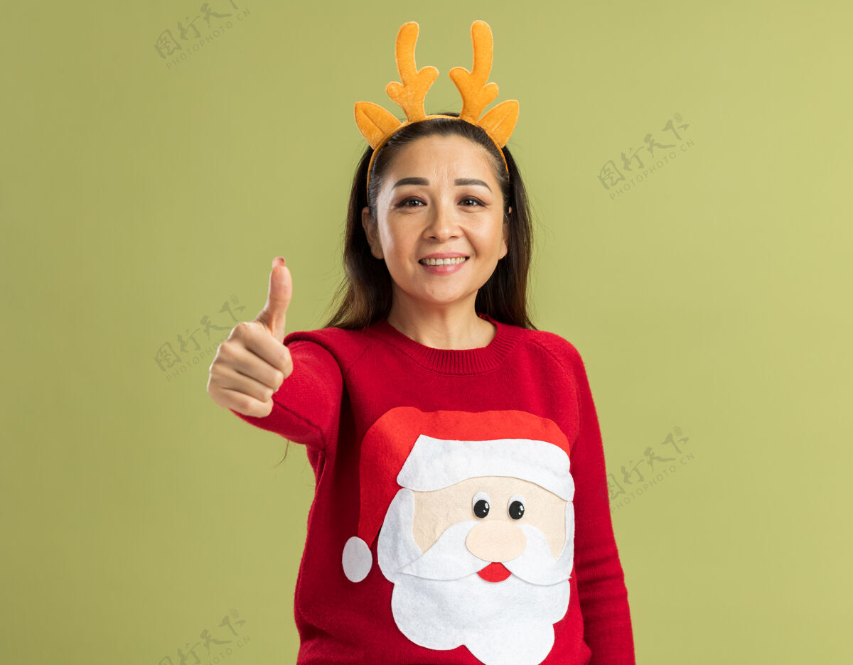 角穿着红色圣诞毛衣的年轻女子 戴着滑稽的鹿角边 微笑着竖起大拇指 快乐而积极女人毛衣向上