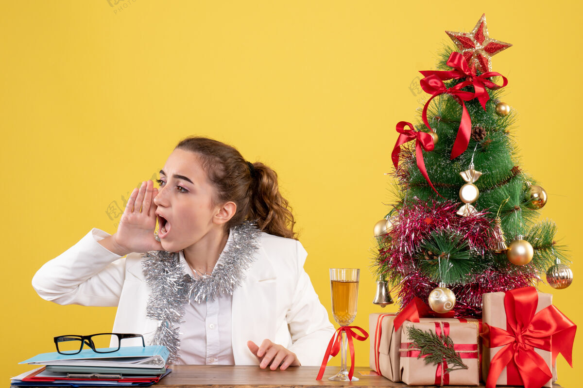 坐着前视图：女医生坐在桌子后面 拿着圣诞礼物 黄色背景上的树在呼唤微笑桌子快乐