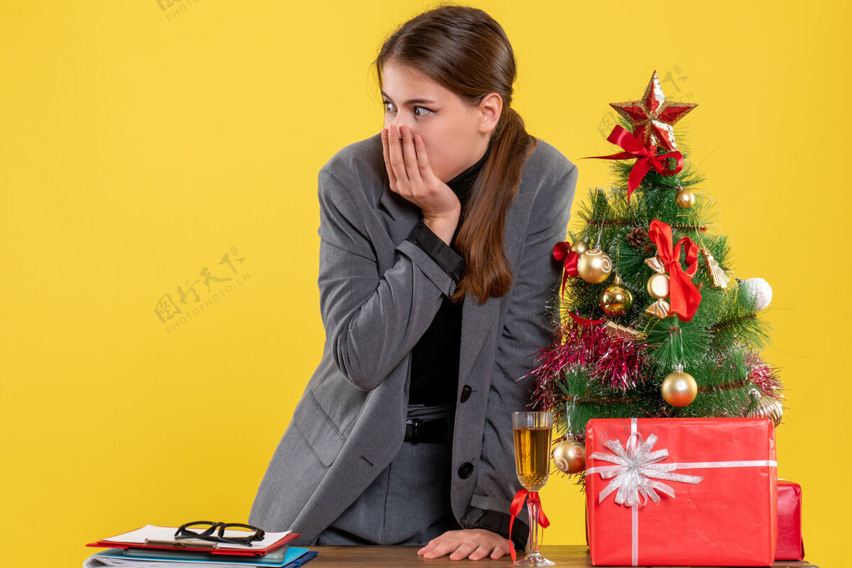圣诞节前视图惊讶的女孩站在桌子后面看着圣诞树和礼物鸡尾酒电脑人惊讶的女孩