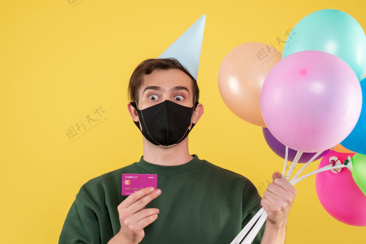帽子正面图：戴着派对帽 戴着黑色面具 手持卡片和气球的年轻人卡片年轻人工具